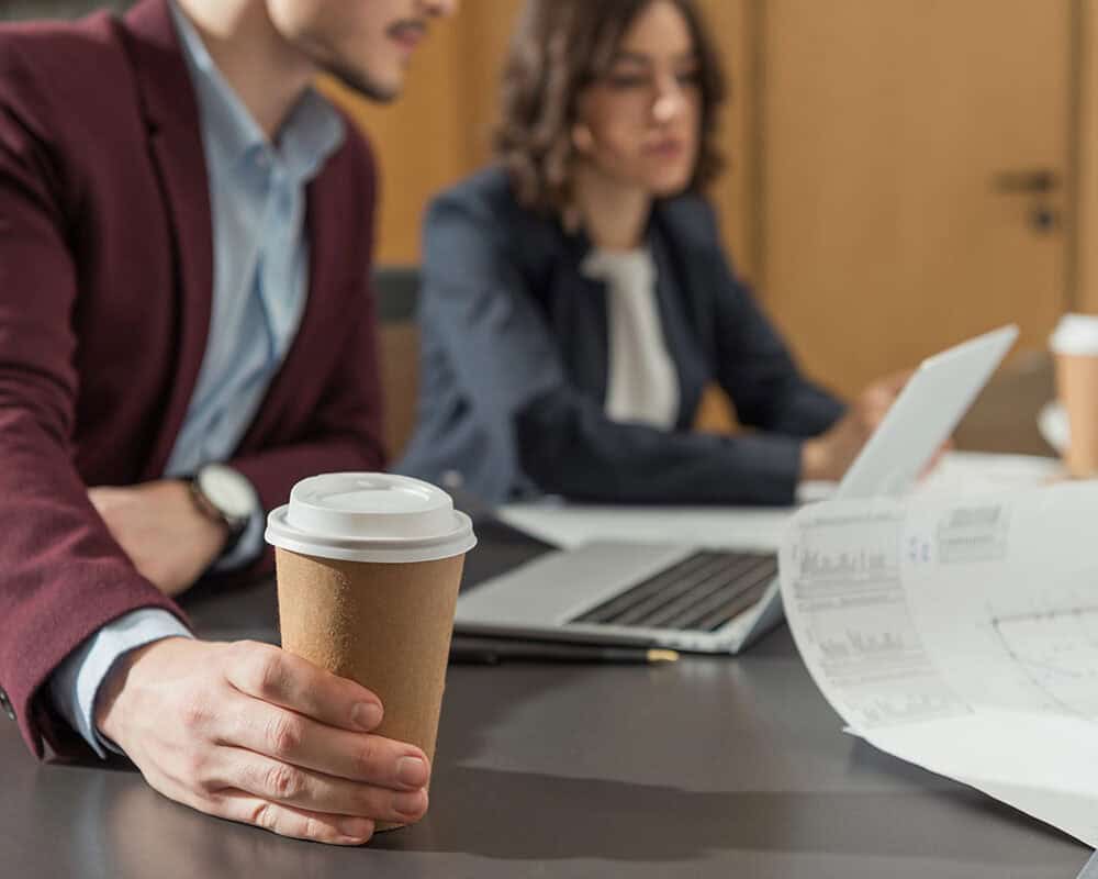 A man in a maroon blazer holds a coffee cup while sitting at a conference table, with a woman in a business suit working on a laptop in the background. Blueprints and business documents are spread across the table.