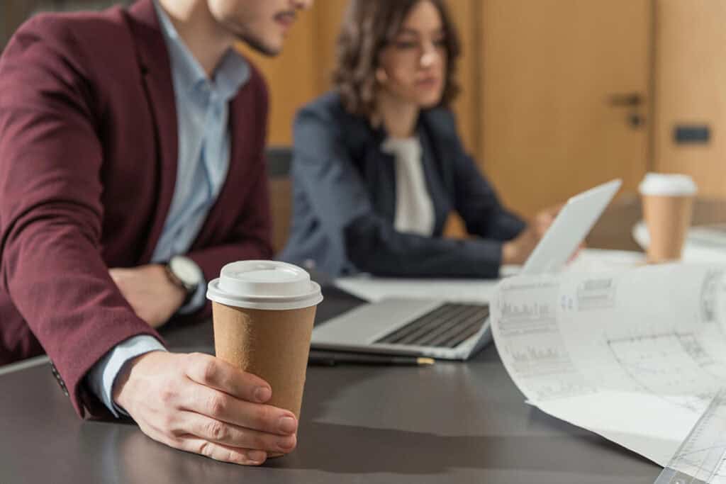 A man in a maroon blazer holds a coffee cup while sitting at a conference table, with a woman in a business suit working on a laptop in the background. Blueprints and business documents are spread across the table.