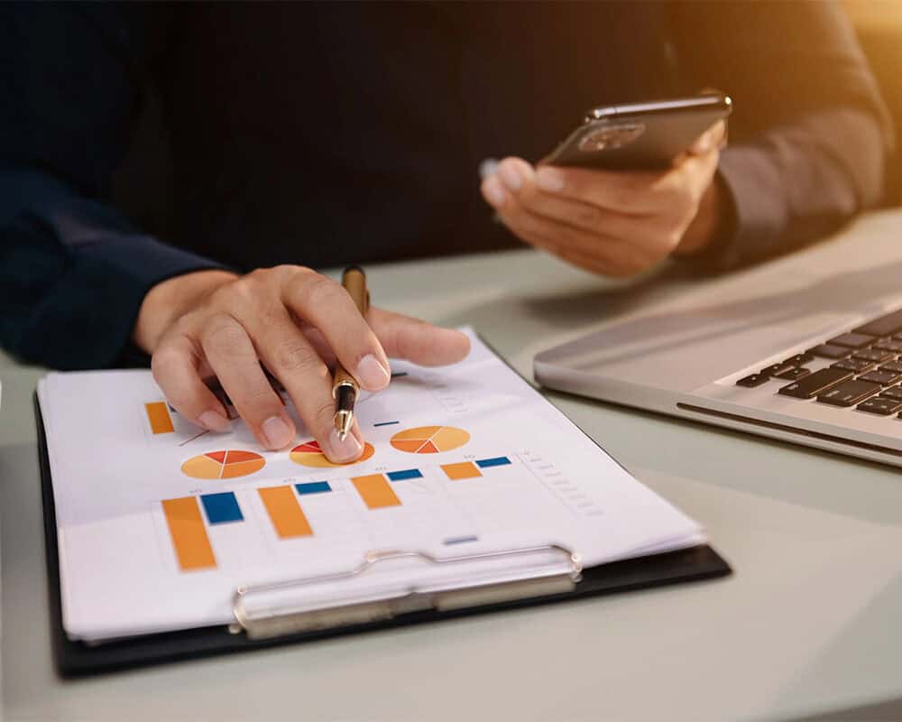 A person's hand holding a pen points at a printed document featuring colorful charts and graphs, while the other hand holds a smartphone. A laptop is visible on the desk alongside a stack of papers.