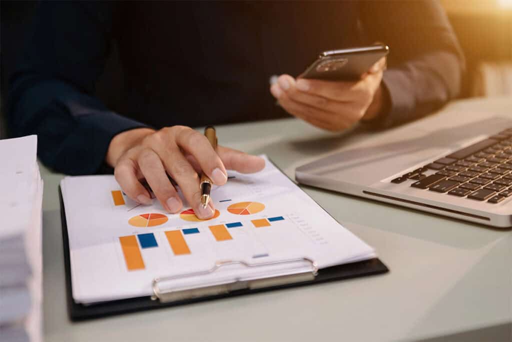 A person's hand holding a pen points at a printed document featuring colorful charts and graphs, while the other hand holds a smartphone. A laptop is visible on the desk alongside a stack of papers.