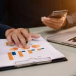 A person's hand holding a pen points at a printed document featuring colorful charts and graphs, while the other hand holds a smartphone. A laptop is visible on the desk alongside a stack of papers.