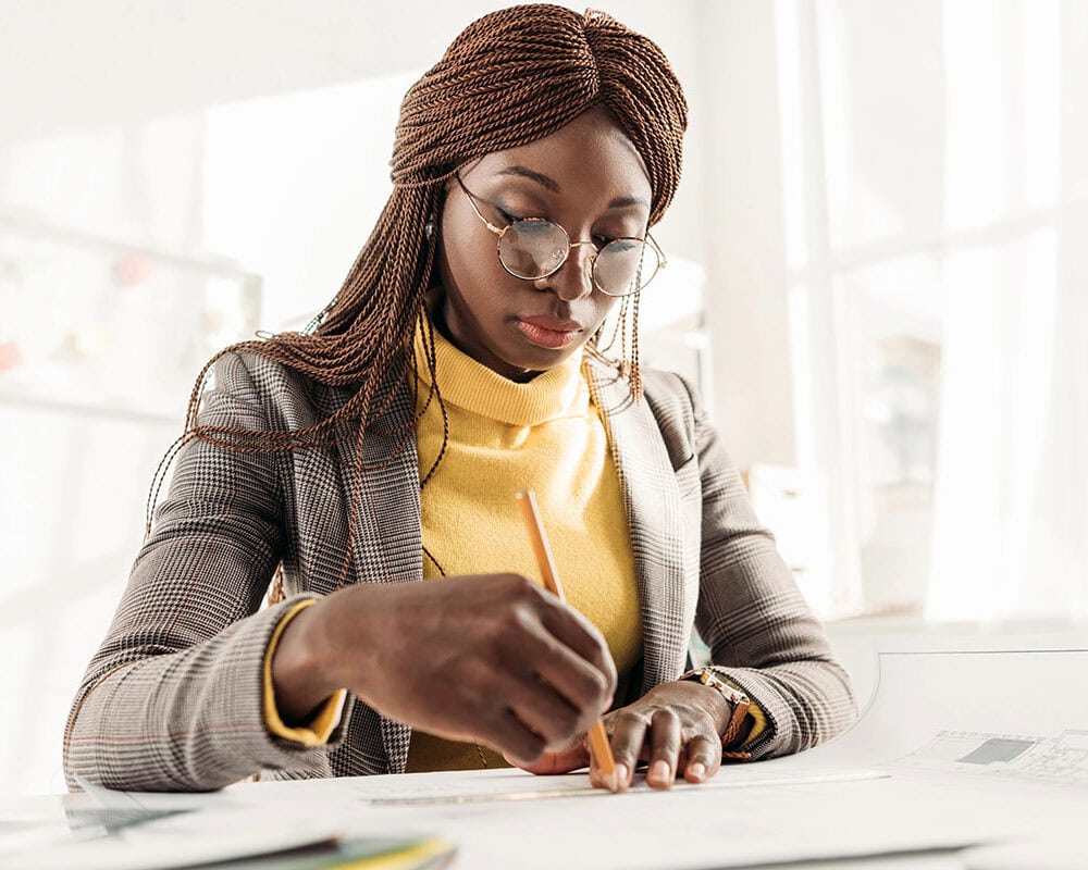 A woman with braided hair, wearing glasses, a plaid blazer, and a yellow sweater, is focused on writing notes with a pencil on a piece of paper at a bright, well-lit workspace.