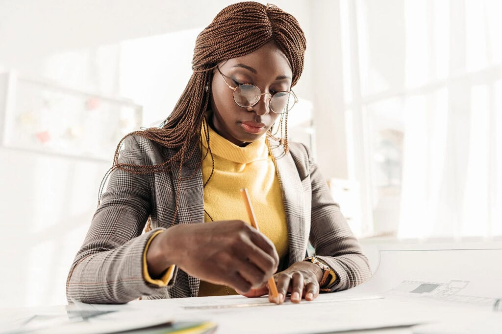 A woman with braided hair, wearing glasses, a plaid blazer, and a yellow sweater, is focused on writing notes with a pencil on a piece of paper at a bright, well-lit workspace.