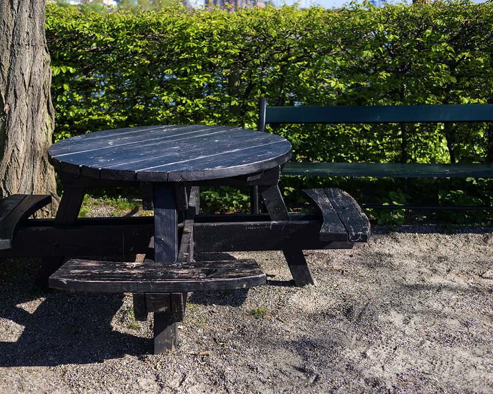 A round black picnic table with attached benches sits on a gravel surface beside a green park bench, surrounded by lush green bushes and trees under a bright blue sky.
