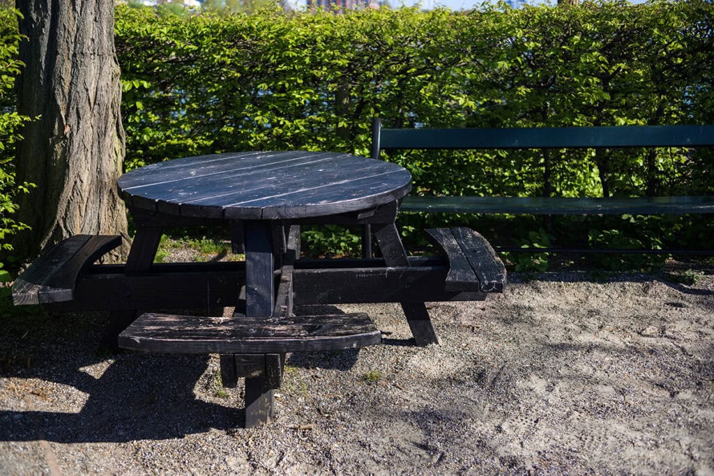 A round black picnic table with attached benches sits on a gravel surface beside a green park bench, surrounded by lush green bushes and trees under a bright blue sky.