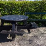 A round black picnic table with attached benches sits on a gravel surface beside a green park bench, surrounded by lush green bushes and trees under a bright blue sky.