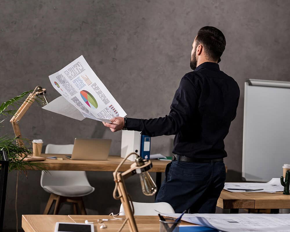 A man in a black shirt stands in an office, holding printed documents with graphs and data. Behind him is a wooden desk with a laptop, stationery, and papers, alongside a plant and a cactus. A whiteboard is positioned in the background.