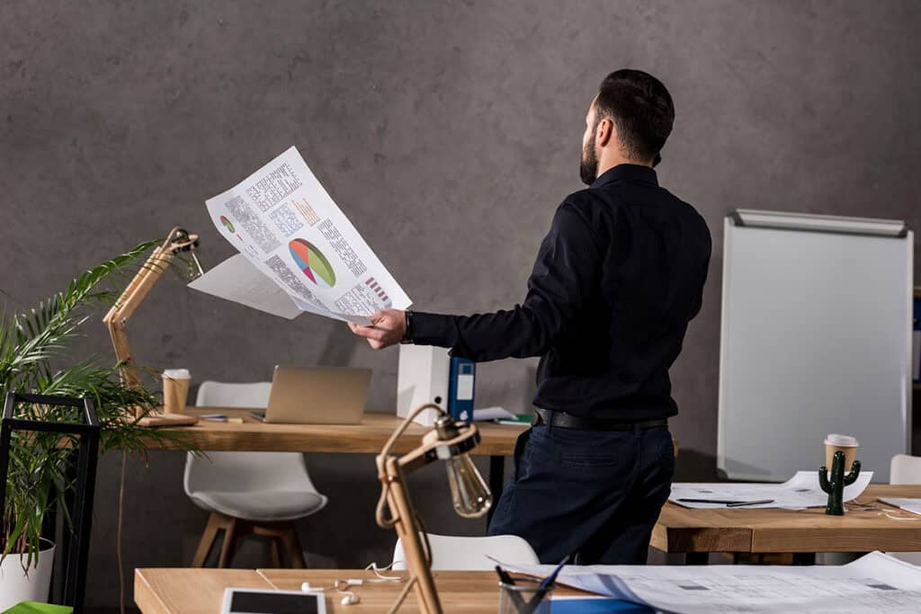A man in a black shirt stands in an office, holding printed documents with graphs and data. Behind him is a wooden desk with a laptop, stationery, and papers, alongside a plant and a cactus. A whiteboard is positioned in the background.
