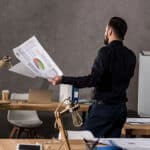 A man in a black shirt stands in an office, holding printed documents with graphs and data. Behind him is a wooden desk with a laptop, stationery, and papers, alongside a plant and a cactus. A whiteboard is positioned in the background.