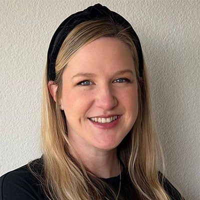 A woman with long blonde hair and a black headband smiles while sitting against a light-colored wall, wearing a black long-sleeve shirt.