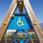 A wooden playground structure featuring a blue sign with a yellow wheelchair symbol, indicating accessibility for individuals with disabilities, against a clear blue sky.