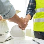 Two individuals shake hands over a table with a laptop, coffee cup, and construction plans. One person wears a reflective yellow safety vest, while the other has a rolled blueprint beside them. A third person is partially visible in the background, holding a clipboard.
