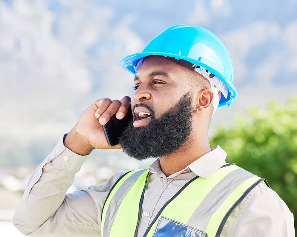 A construction worker wearing a blue hard hat and a reflective safety vest is speaking on a smartphone outdoors, with mountains and greenery in the background.