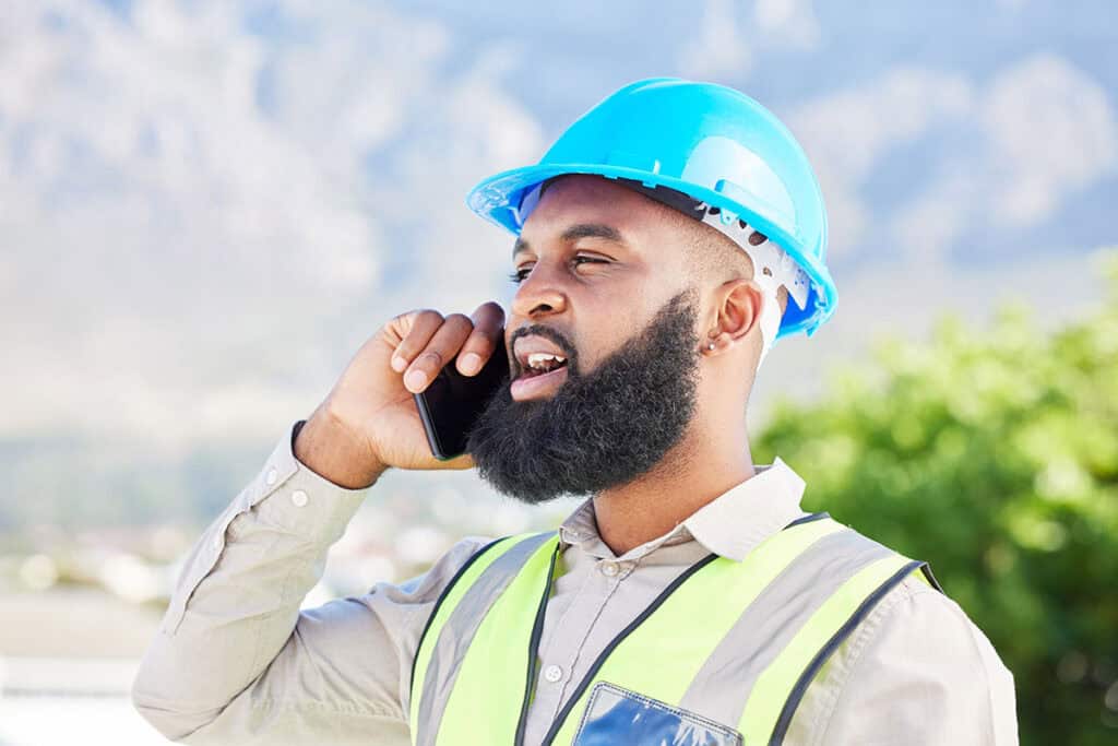 A construction worker wearing a blue hard hat and a reflective safety vest is speaking on a smartphone outdoors, with mountains and greenery in the background.