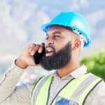 A construction worker wearing a blue hard hat and a reflective safety vest is speaking on a smartphone outdoors, with mountains and greenery in the background.