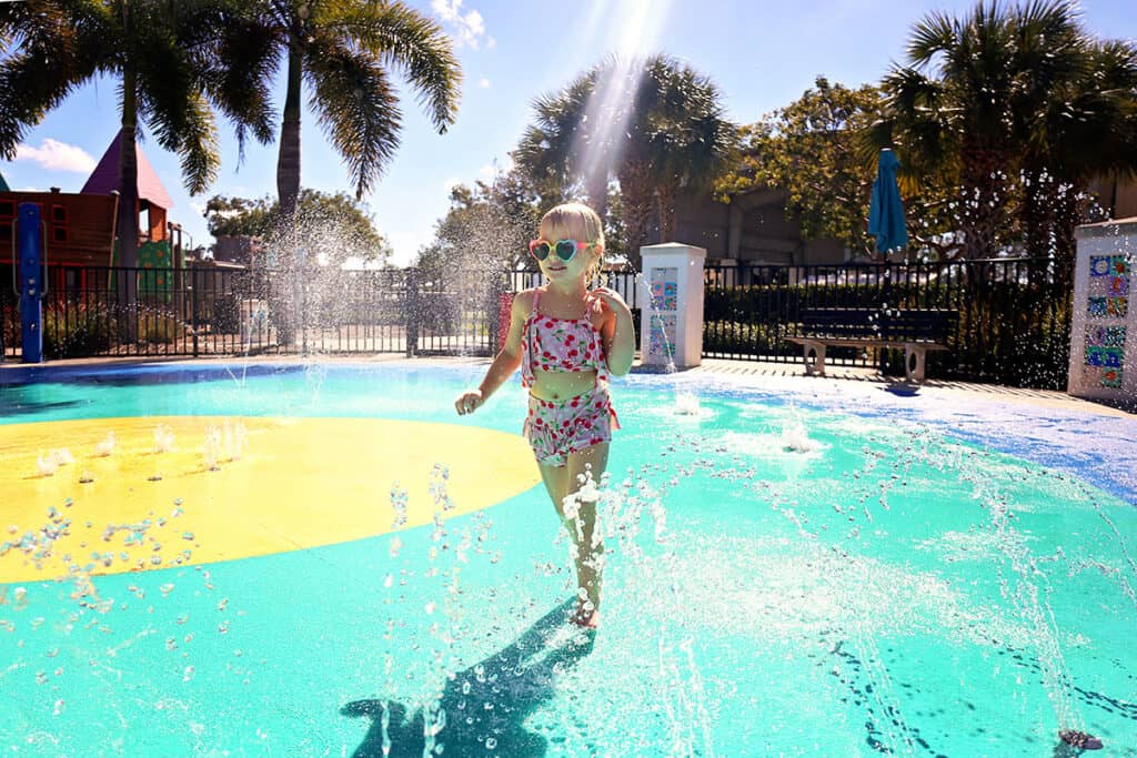 A young girl with short blonde hair and heart-shaped sunglasses joyfully runs through a splash pad, with colorful water jets spraying around her. Palm trees and a play structure can be seen in the background under a bright blue sky.