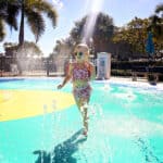 A young girl with short blonde hair and heart-shaped sunglasses joyfully runs through a splash pad, with colorful water jets spraying around her. Palm trees and a play structure can be seen in the background under a bright blue sky.