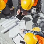 An overhead view of three construction workers wearing hard hats and high-visibility vests gathered around architectural blueprints and a digital tablet on a concrete floor.