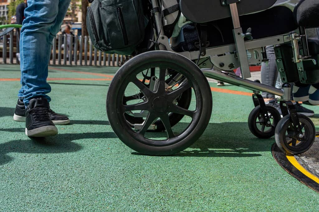 A low-angle view of a wheelchair wheel and a person standing on a green, padded playground surface next to an accessible roundabout ride.