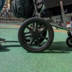 A low-angle view of a wheelchair wheel and a person standing on a green, padded playground surface next to an accessible roundabout ride.