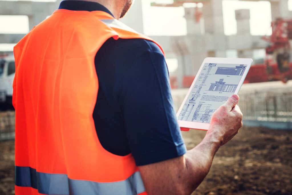 A construction worker wearing a high-visibility vest reviews digital building plans on a tablet at an active job site.