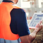 A construction worker wearing a high-visibility vest reviews digital building plans on a tablet at an active job site.