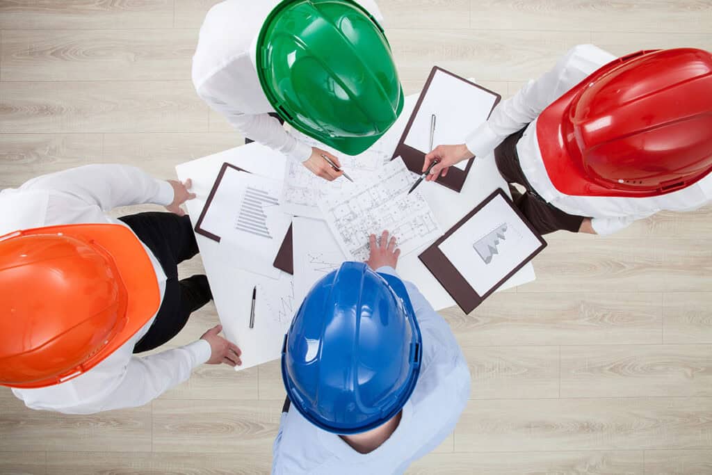 Overhead view of four professionals in colored hardhats gathered around a table reviewing blueprints and financial charts.
