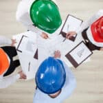 Overhead view of four professionals in colored hardhats gathered around a table reviewing blueprints and financial charts.