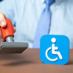 A close-up of a person in business attire raising a red rubber stamp beside a blue International Symbol of Access icon on a desk.