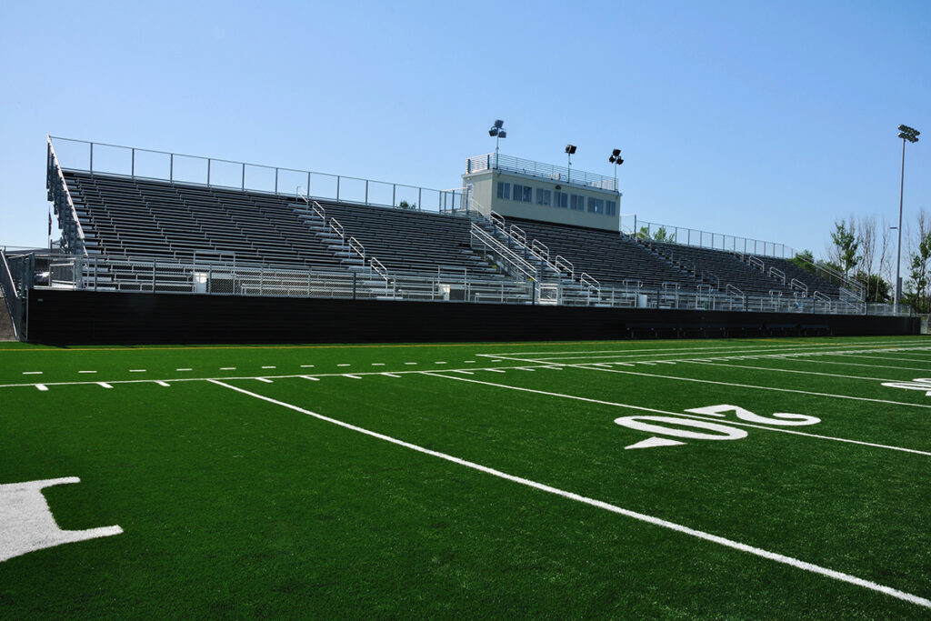 Empty stadium bleachers and a press box bordering a green football field with yard line markings under a clear blue sky.