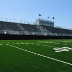 Empty stadium bleachers and a press box bordering a green football field with yard line markings under a clear blue sky.