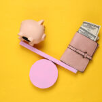 A top-down view of a pink seesaw balancing a pink piggy bank on the left and a leather wallet filled with US dollar bills on the right, set on a yellow surface.